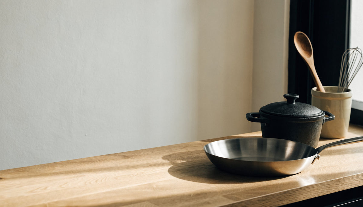 Poêle en inox, cocotte en fonte et ustensiles en bois sur un plan de travail en chêne
