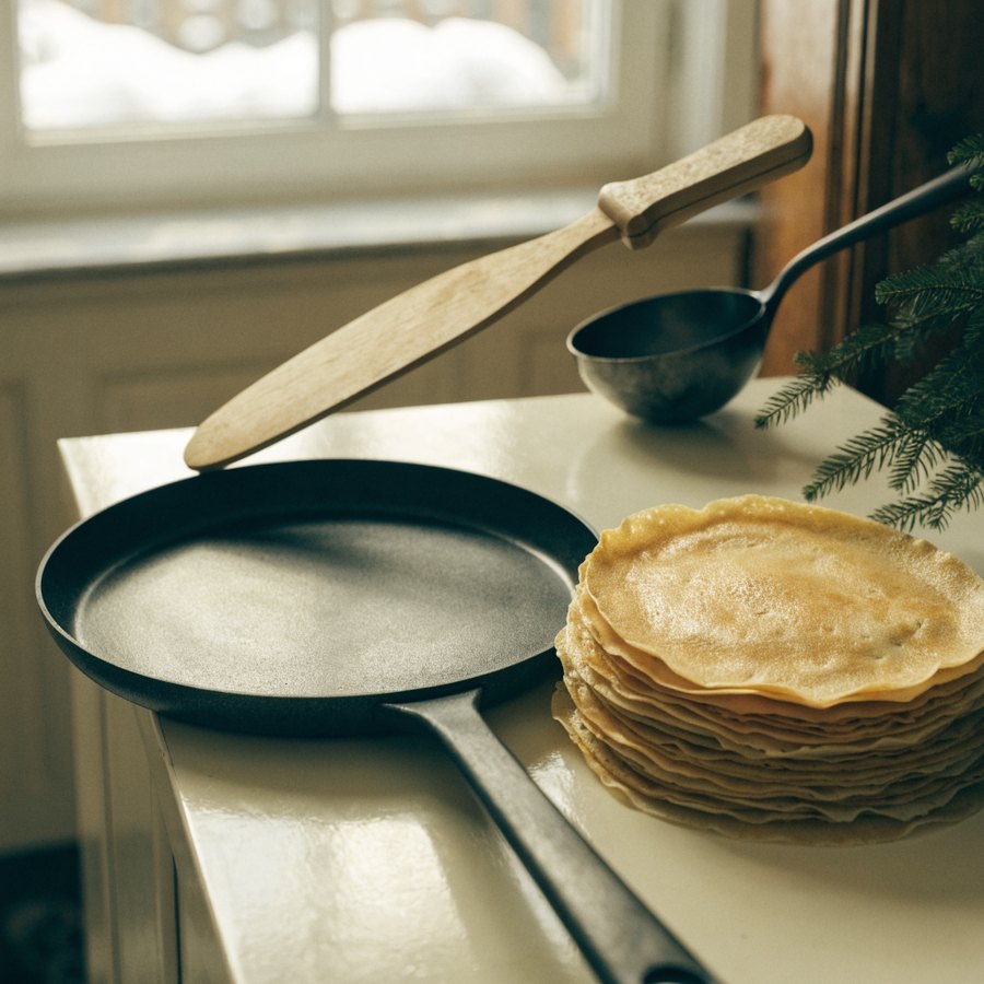 Crêpière en fonte, pile de crêpes et râteau en bois devant une fenêtre d'hiver